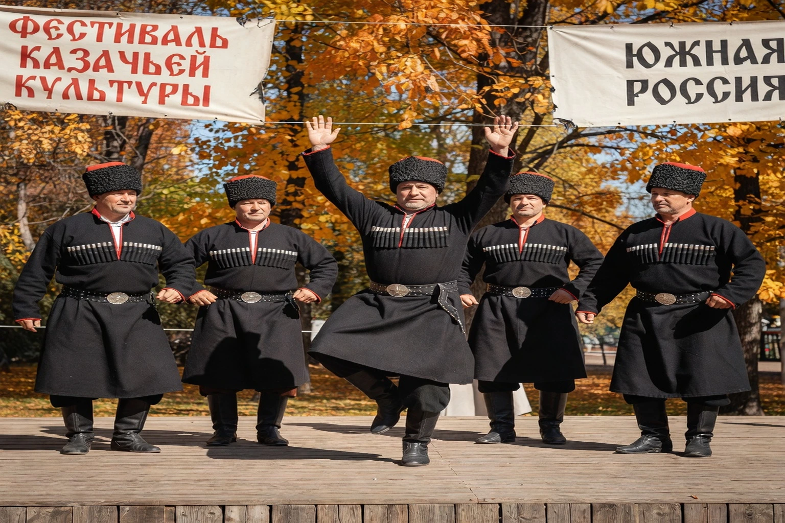 Ensemble folklorique cosaque de cinq hommes en tcherkeska noire dansant sur une scène en plein air, coiffés de kubankas d'astrakan noir à calot rouge